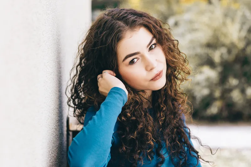 Woman with curly hair in a blue sweater leaning against a wall and looking at the camera