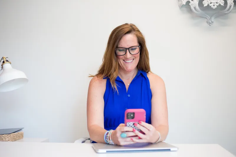 Smiling woman in a blue top using a pink smartphone at her desk