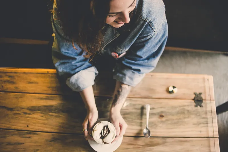Woman laughing at a wooden café table with a cappuccino, seen from above