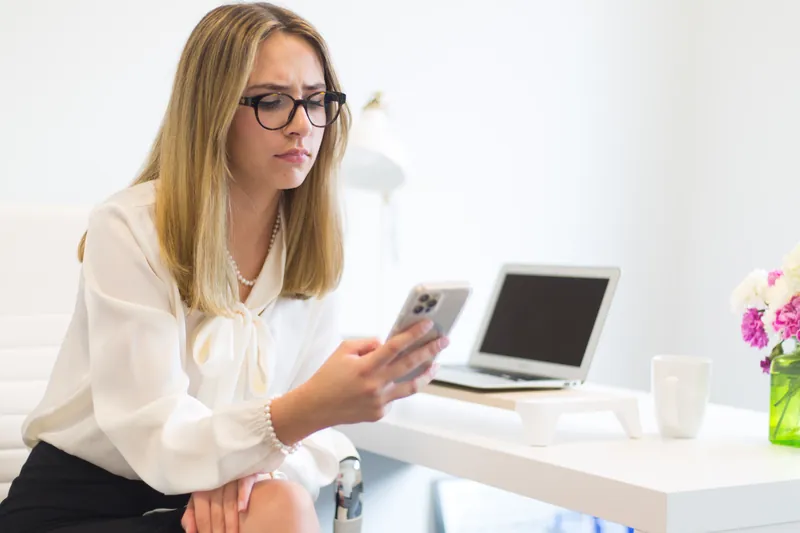 Woman with glasses checking her phone at a bright desk with a laptop