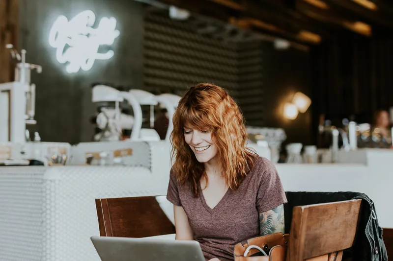 Red-haired woman laughing while using a laptop in a café