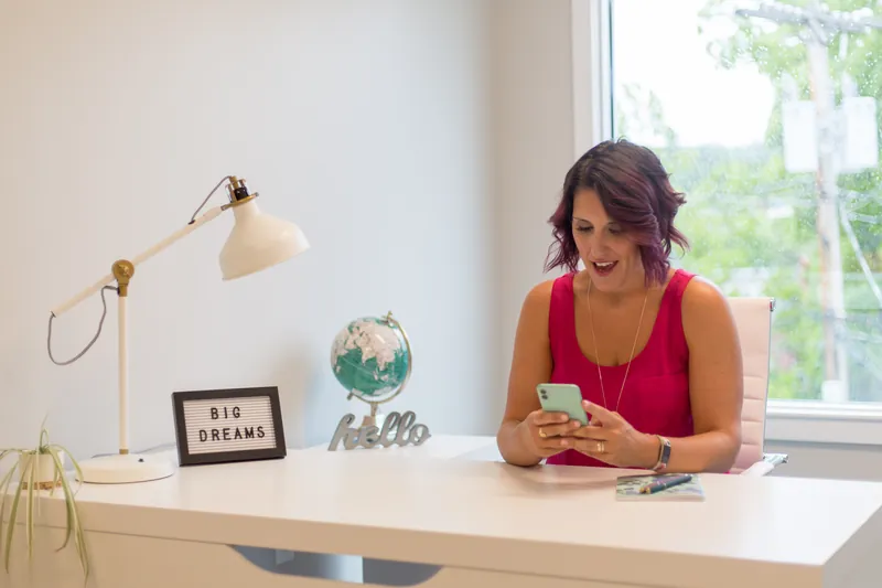 Woman in a pink top laughing at her phone at a bright desk