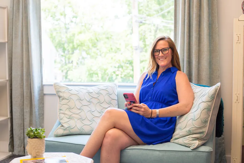 Smiling woman in a blue dress sitting on a sofa with her smartphone