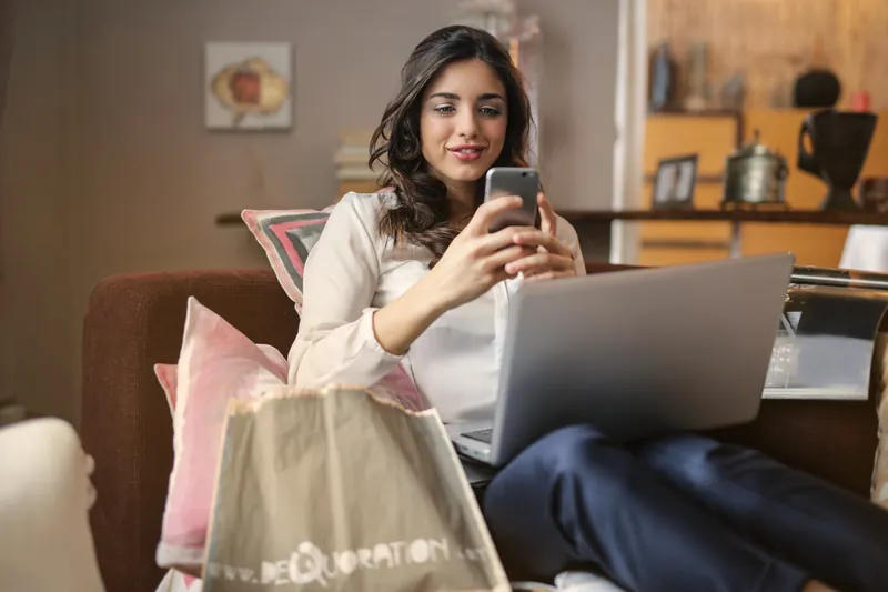 Woman on a sofa smiling at her phone with a laptop on her lap